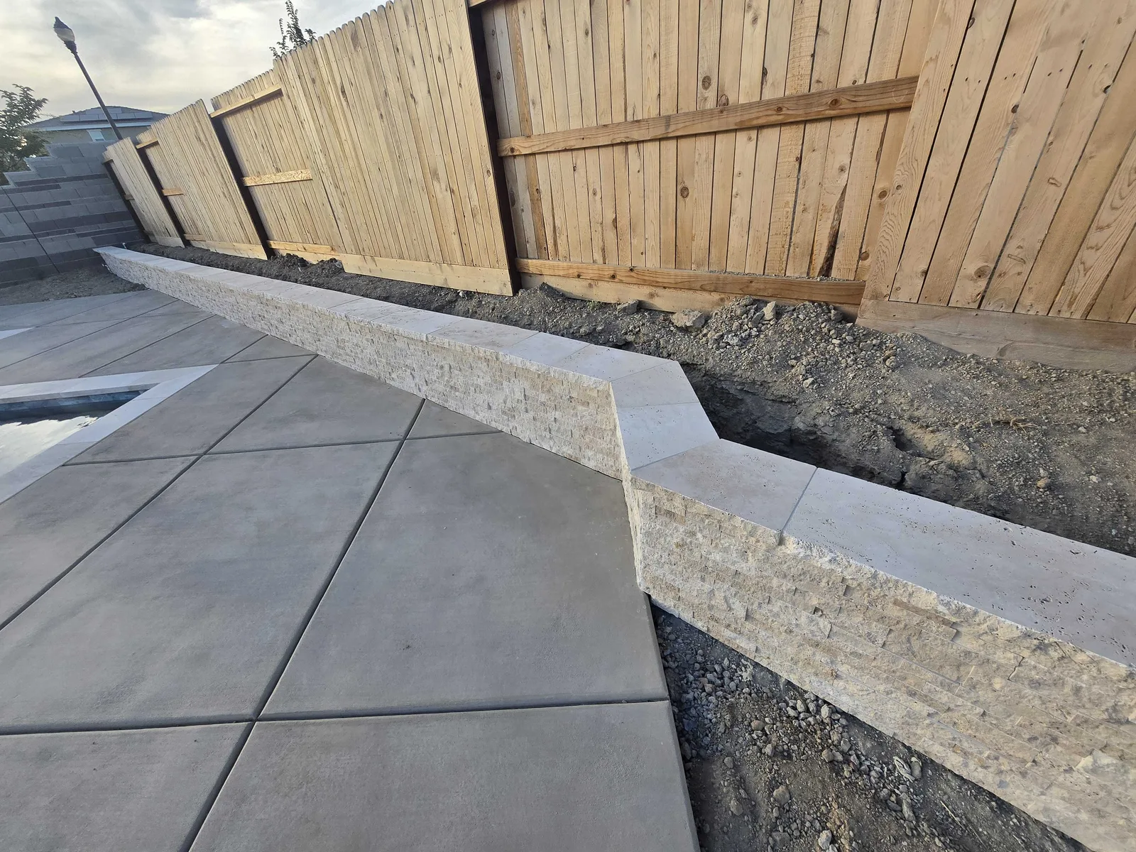 Stone-veneer block retaining wall with a cap along a fence line beside a new concrete pool deck