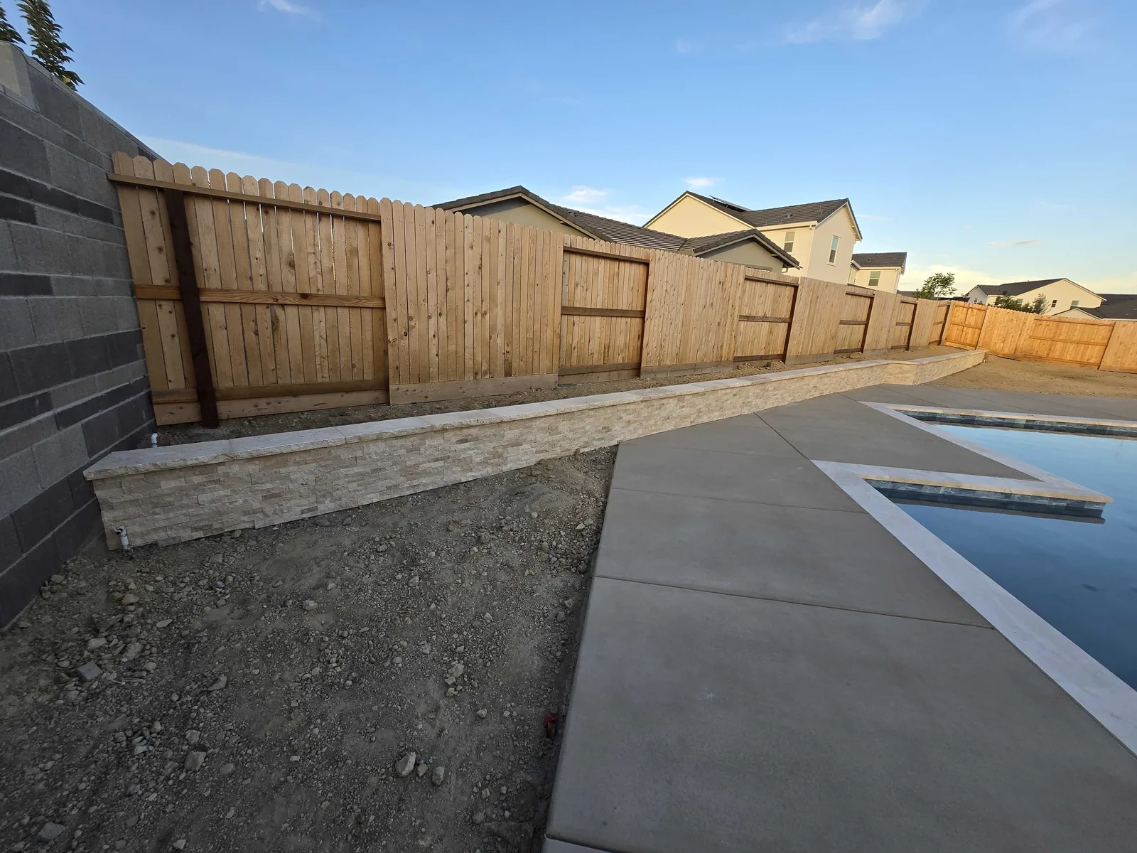 Low stone-veneer retaining wall with a cap running along a fence line beside a new concrete pool deck