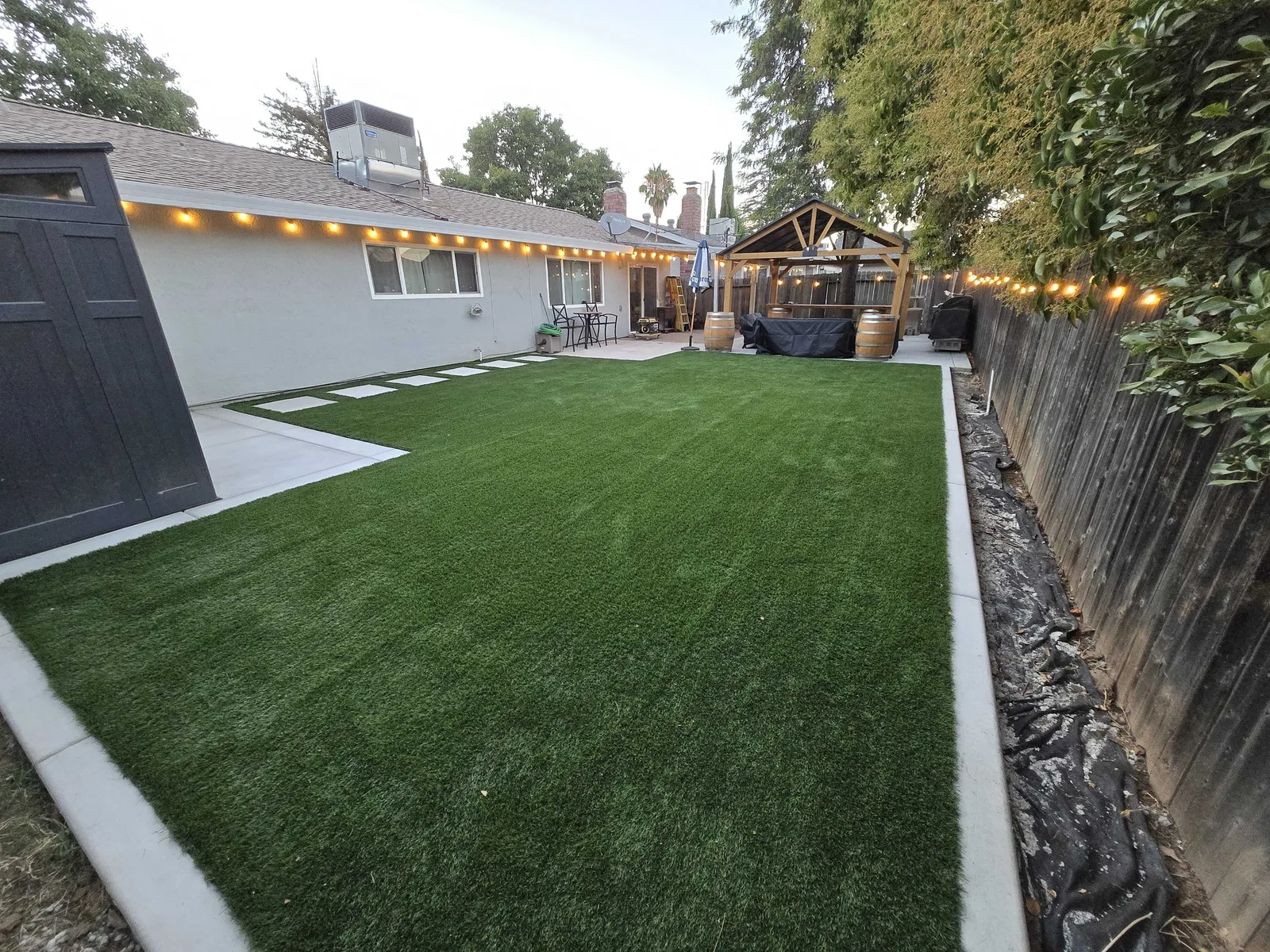 Backyard artificial turf lawn edged by a concrete patio and concrete stepping pads, with string lights along the fence and a covered pavilion behind